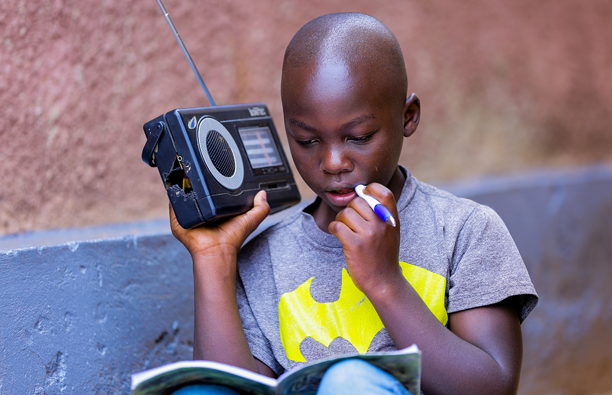 A young boy in Rwanda holds up a radio to his ear to hear his lessons during school closures. He has an exercise book on his knee and a pen in his hand.
