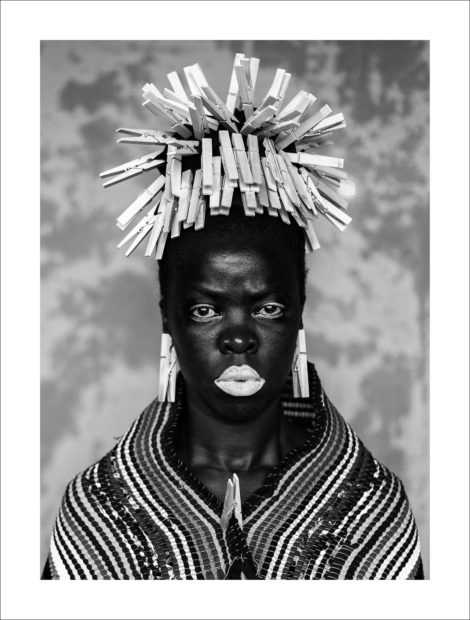 Self-portrait by Zanele Muholi with clothes pegs in their hair and on her ear lobes and wearing white lipstick.
