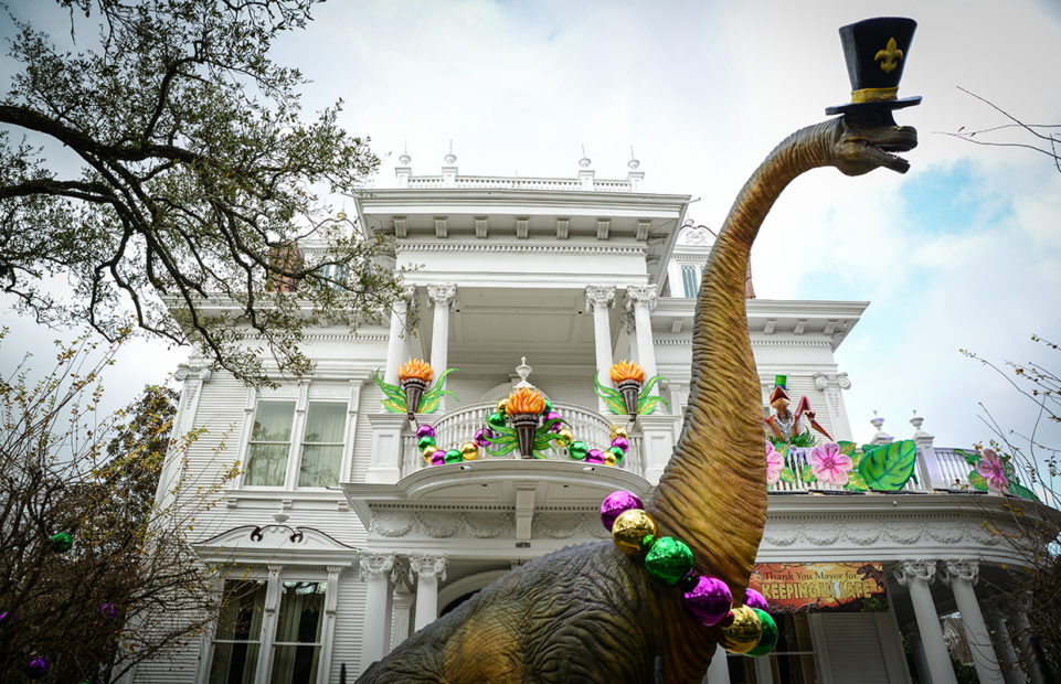 A house decorated as a Mardi Gras float in New Orleans. A long-nicked dinosaur sports a top hat and tradition Mardi Gras beads in the traditional colours: purple, gold and green.