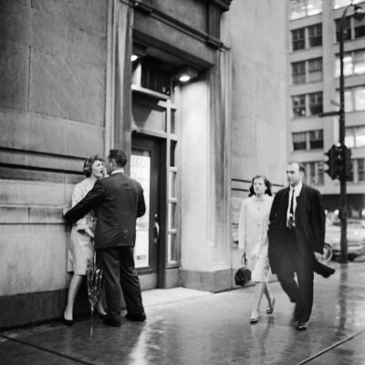 Street scene with one couple walking peacefully and another behind: the man has the woman pushed against the wall of a building