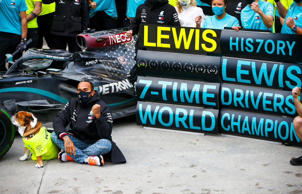 Lewis Hamilton with a mask seated beside a dog in front of a racing car a display reading: Lewis History Lewis 7-time drivers' world champion