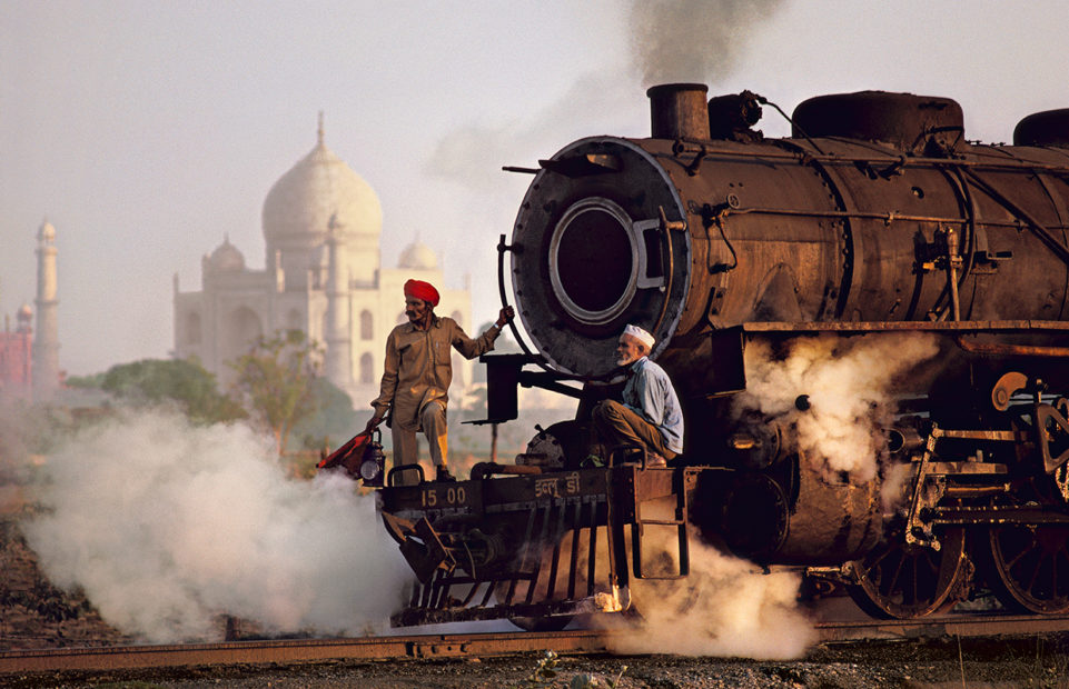 Two men stand on teh front of the engine of a steam train in Uttar Pradesh, India, 1983, with the Taj Mahal n the background.