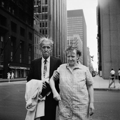 An elderly man and woman, rather disheveled, walking in the middle of a city street surrounded by skyscrapers.
