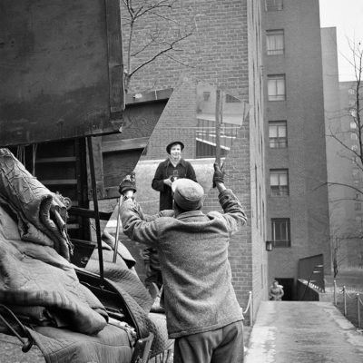 Vivian Maier reflected in a mirror held by a man who seems to have lifted it from a pile of discarded objects.