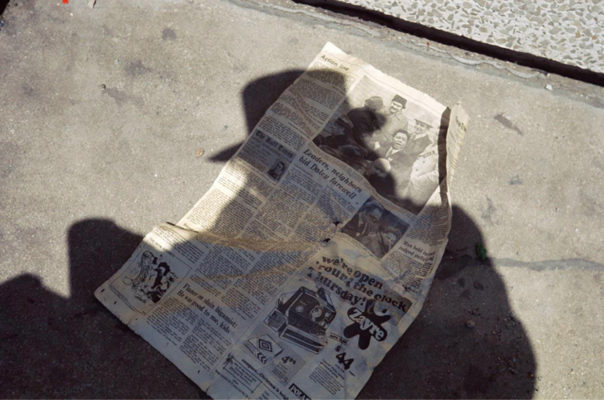 A crumpled newspaper ona sidewalk with a shadow of a person in a hat covering it.