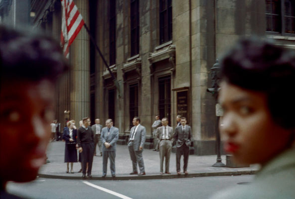 In the foreground, two smartly dressed young black women, looking back at the camera, out of focus. On the other side of the street a group of white men, with an American flag in the background.