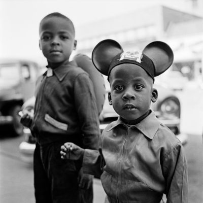 Two African American boys looking at the camera. The smaller on, on the right, is wearing Mickey Mouse ears, the older one has a cowboy hat slung on his back.