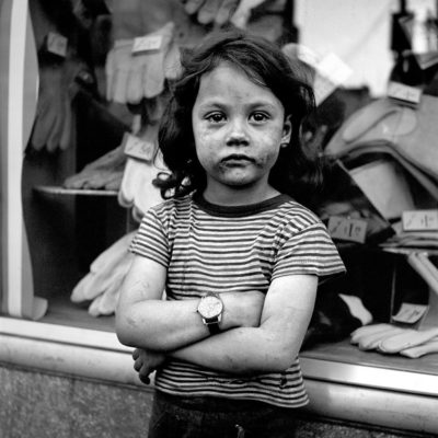 A girl with her arms crossed wearing a stripy T-shirt and a larg watch, outside a shop