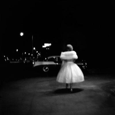A woman in a white evening dress and fur wrap, from behind, outside in the dark.