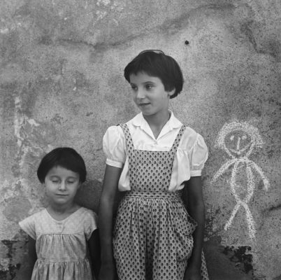 Two little girls standing against a wall, with a chalk drawing of a third child.