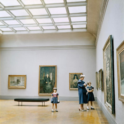 A girl and a woman looking at a painting in a museum. A second girl stands apart, looking at the camera.