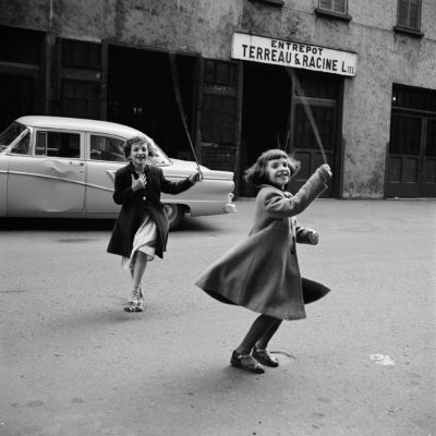 Two young girls playing with a skipping rope in front of a warehouse with a French name.