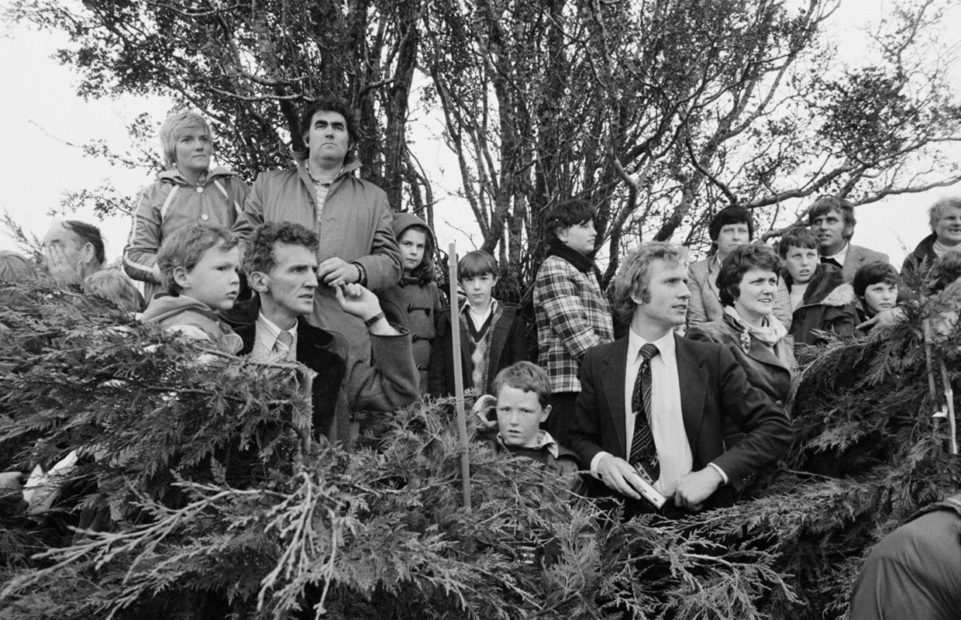 A black and white photo of a group people who seem to be standing in a hedge looking at something.
