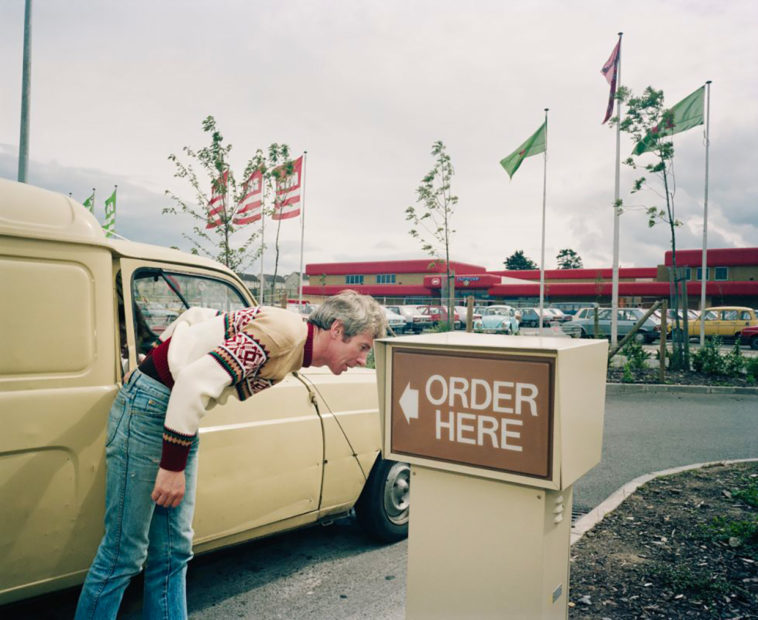 A man who has got out of his car and bent down to speak into the microphone to order drive-through McDonalds.