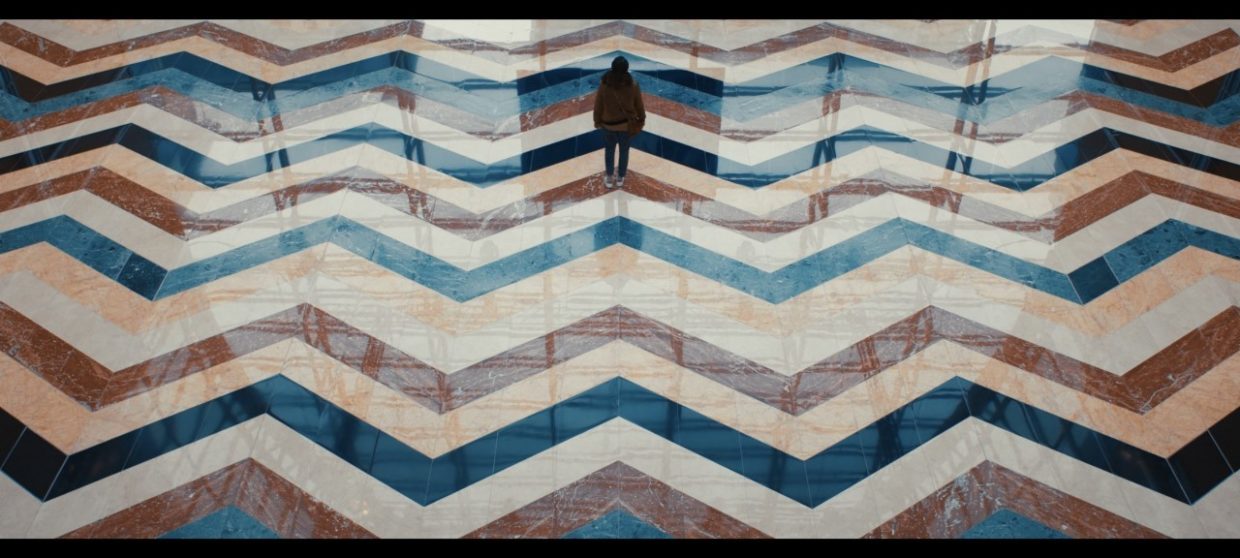 A young woman stands on a vast tile floor covered in reflections