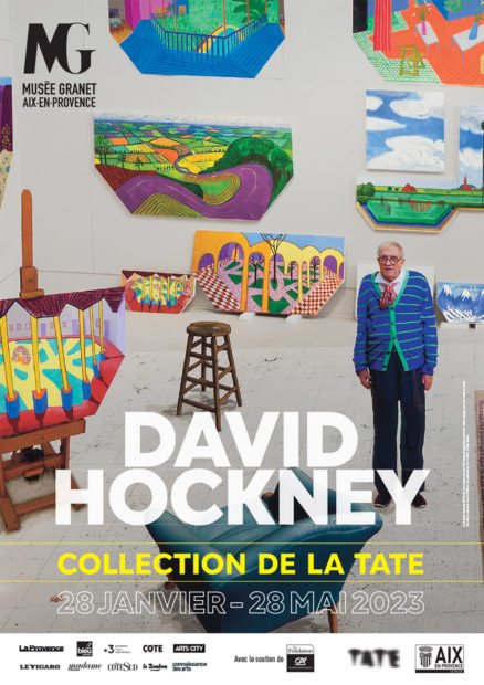 Poster for the exhibition featuring Hockney standing in his studio surrounded by multicoloured images. 