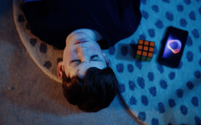 A still from the film Lia IRL. We see a boy lying down next to a Rubik's Cube and a phone.