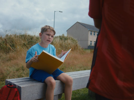 A still from the film Spines. A boy in shorts is reading a larg e book and looking at someone standing on the edge of the frame.