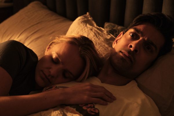 A British Indian man and a white woman in bed. She is sleeping, he is staring into space.