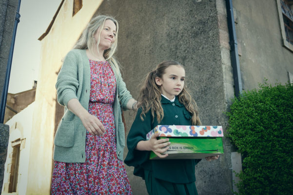 A still from the film Wider than the Sky. A girl is holding abox and walking with her mother, both looking serious.