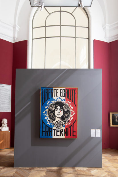 A red, white and blue in the form of the French flag with a woman's face in Art Nouveau woodcut style and the words Liberté, Egalité, Fraternité,. There is a single red tear on the woman's face.