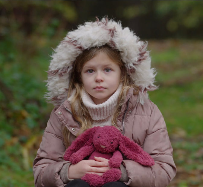 A girl wearing a parka with the hood up and holding a pink toy rabbit, staring at the camera.