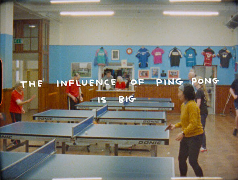 A still of people playing table tennis in a large room with the title of the film superimposed; The Influence of Ping Pong is Big