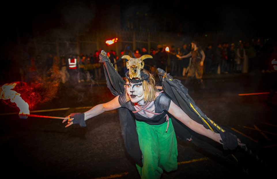A performer in a spooky black and green costume with white and black face paint and an animal's skull as a headpiece, brandishing fiery torches.