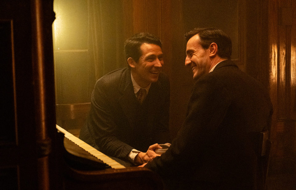 A still from The History of Sound with Josh O'Connor and Paul Mescal laughing next to an upright piano.