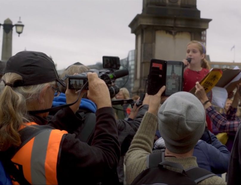 A girl being filmed by the crowd as she gives a speech in the open air.