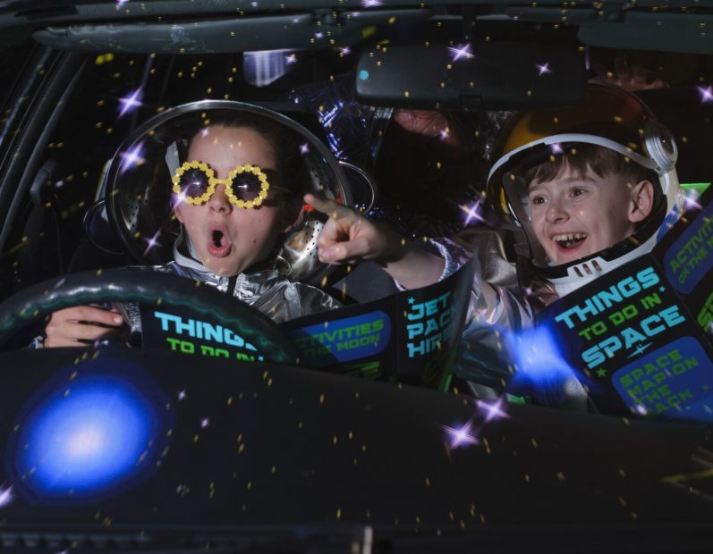 Kids in a car in the dark wearing space suits and reading leaflets entitled Things to do in Space.