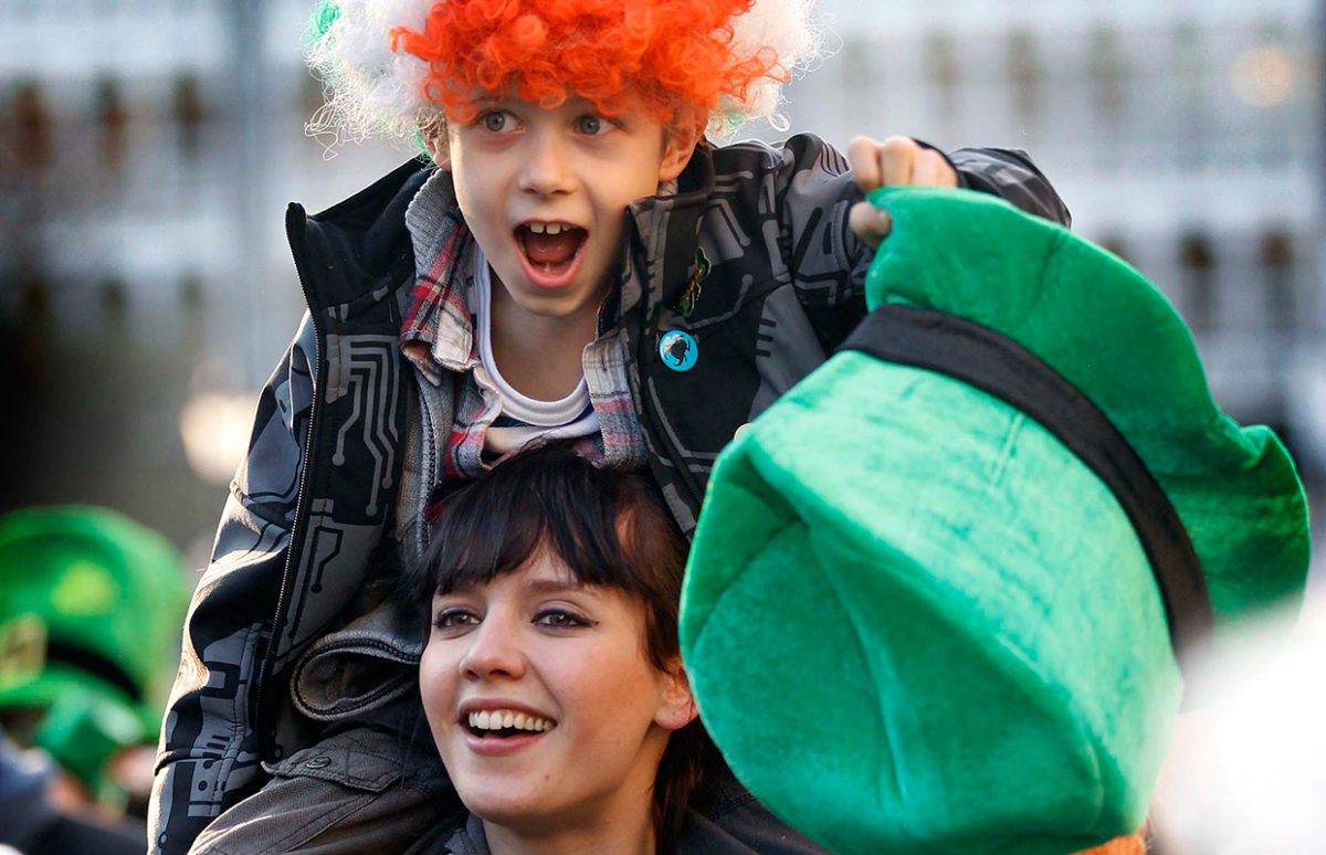 A boy in an Irish tricolour wig sitthing on the shoulders of a young woman and holding a green leprechaun hat