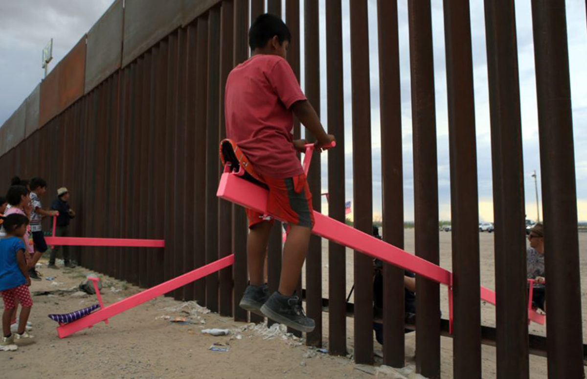 A child on one end of a pink see-saw that is placed through the U.S.-Mexico border wall.