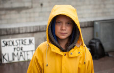 Greta Thunberg in a yellow rain jacket with the hood up. A sign saying in Swedish School Strike for the Planet can be glimpsed in the background.