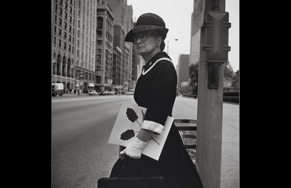 A middle-aged woman about to cross a city street with a painting under her arm.