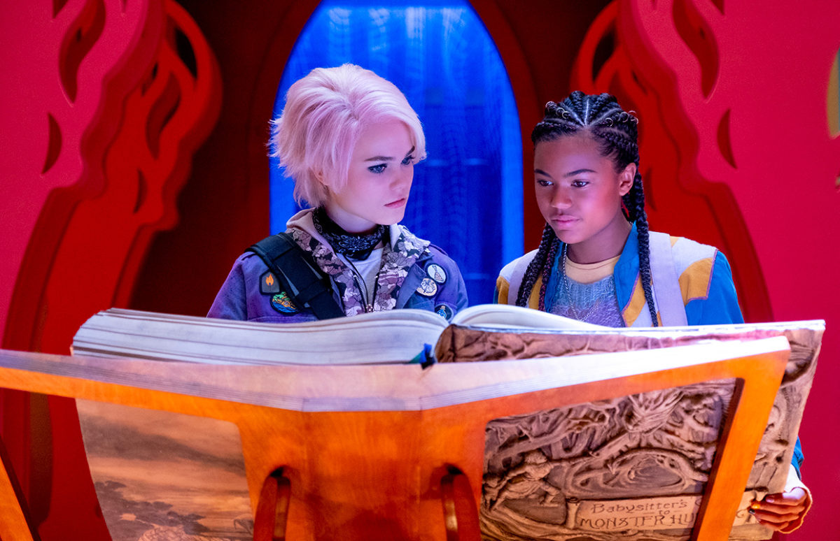 Liz and Kelly, Babysitters from the film, looking at a large book in Babysitters' HQ.