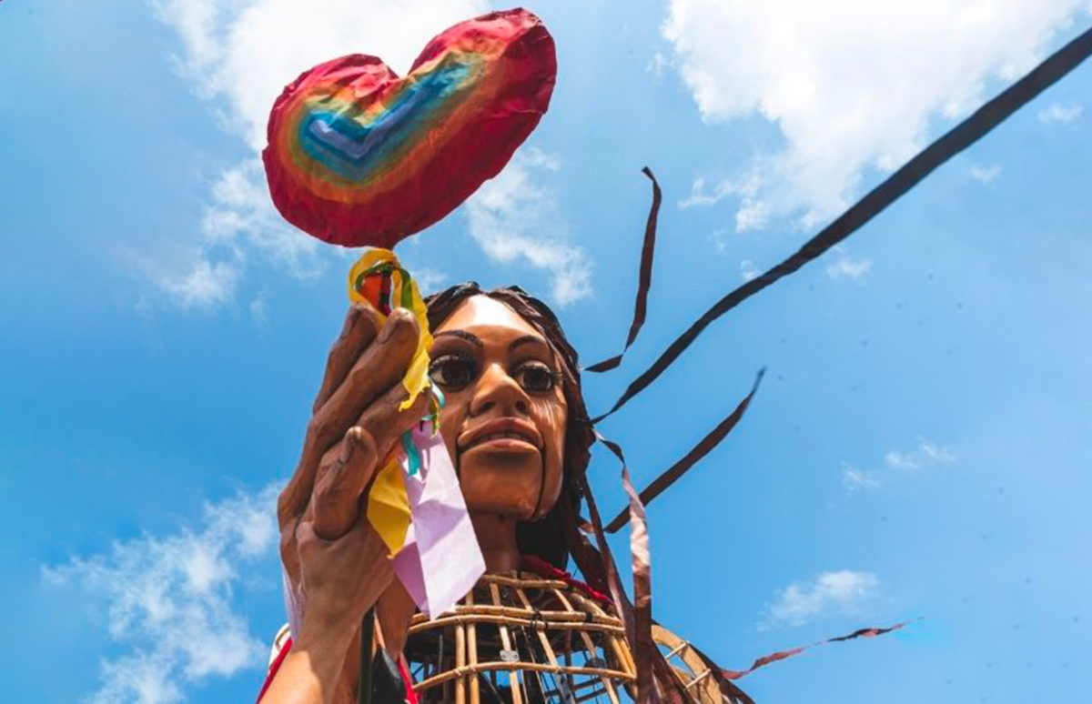 The Little Amal puppet holding a rainbow-coloured heart-shaped balloon.