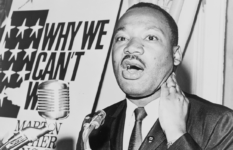 Martin Luther King speaking into a microphone in front of a poster with an American flag.
