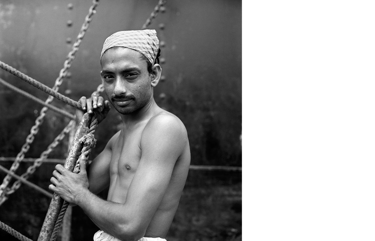 1959. Kochi, India VM1959W04098-13-MC A young man with a bare chest and a bandana covering his hair posing in front of a large ship.