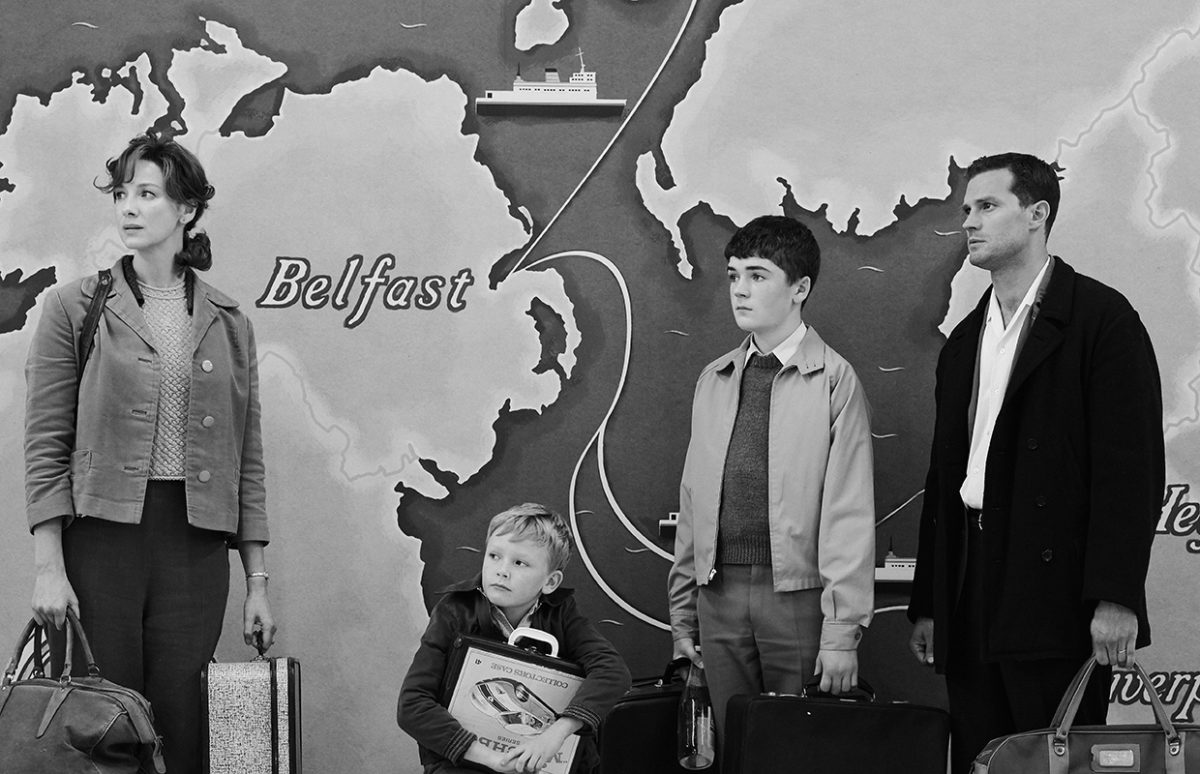 The family in the film Belfast standing with luggage in front of a mural that is a map showing ways to leave Northern Ireland