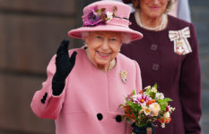 Her Majesty the Queen dressed in pink, waving and carrying a bouquet of flowers.
