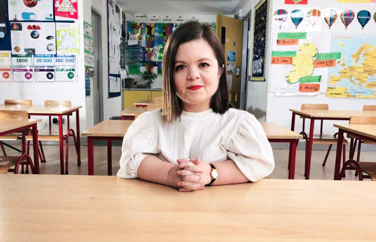 Sinéad Burke sitting at a desk in an empty classroom. There is a map of Ireland on the wall behind her.