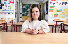 Sinéad Burke sitting at a desk in an empty classroom. There is a map of Ireland on the wall behind her.