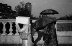 A black-and-white photo of people walking in the rain with umbrell's. One woman has a cardboard box protecting her head.