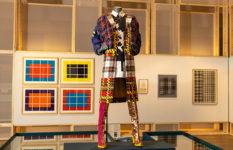 A mannequin displaying an outfit in many different tartans, with more tartan patterns framed on the wall in the background.