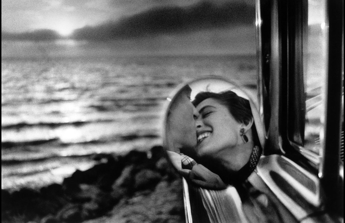 A black and white photo of a couple glimpsed in the wing mirror of their car, parked by the ocean