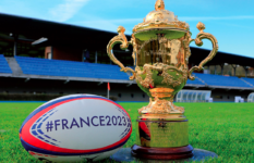 The Webb Ellis World Cup and a rugby ball reading France2023 on the grass of a stadium.