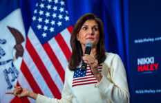 NIkki Haley holding a microphone standing in front of an American flag and with another on her jumper.