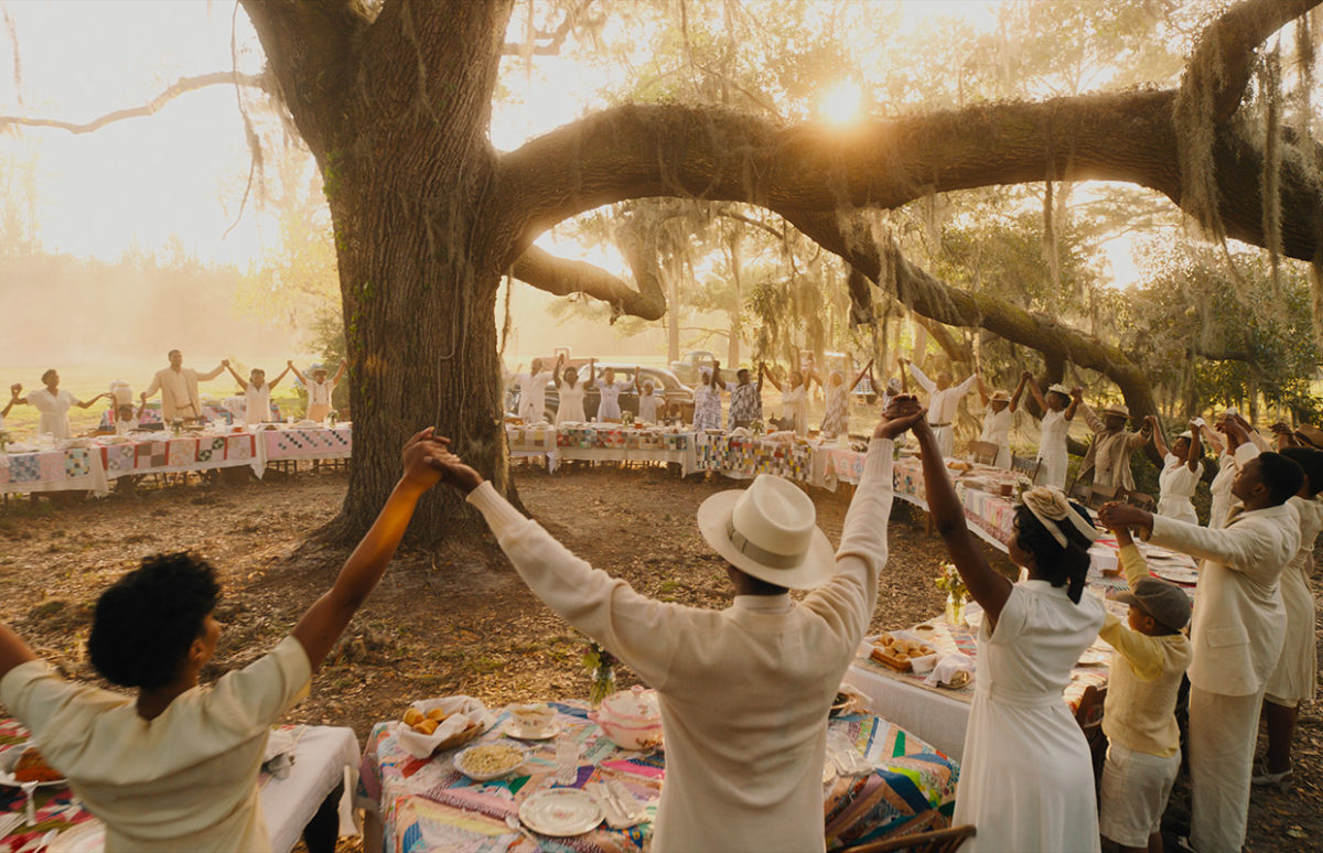 A large group of African Americans holding hands at picnic tables set around a large tree.