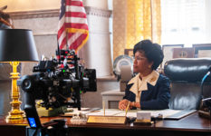 Regina King as Shirley Chisholm, being filmed at a desk with an American flag in the background.